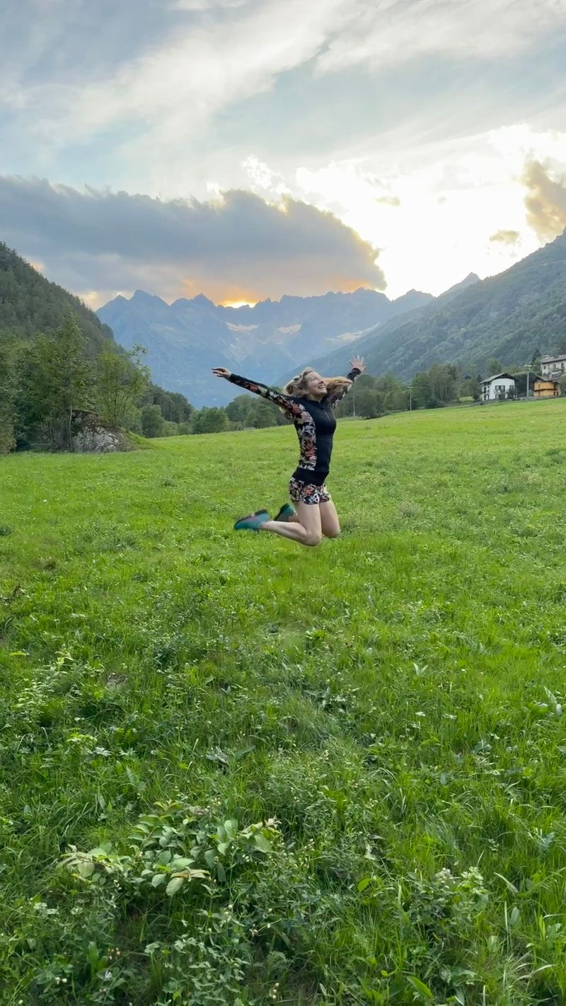 A woman jumping for joy in a green meadow below mountains at sunset